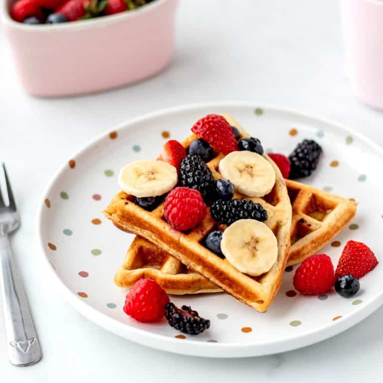 3-ingredient protein waffles stacked on a plate topped with fruit and bananas.