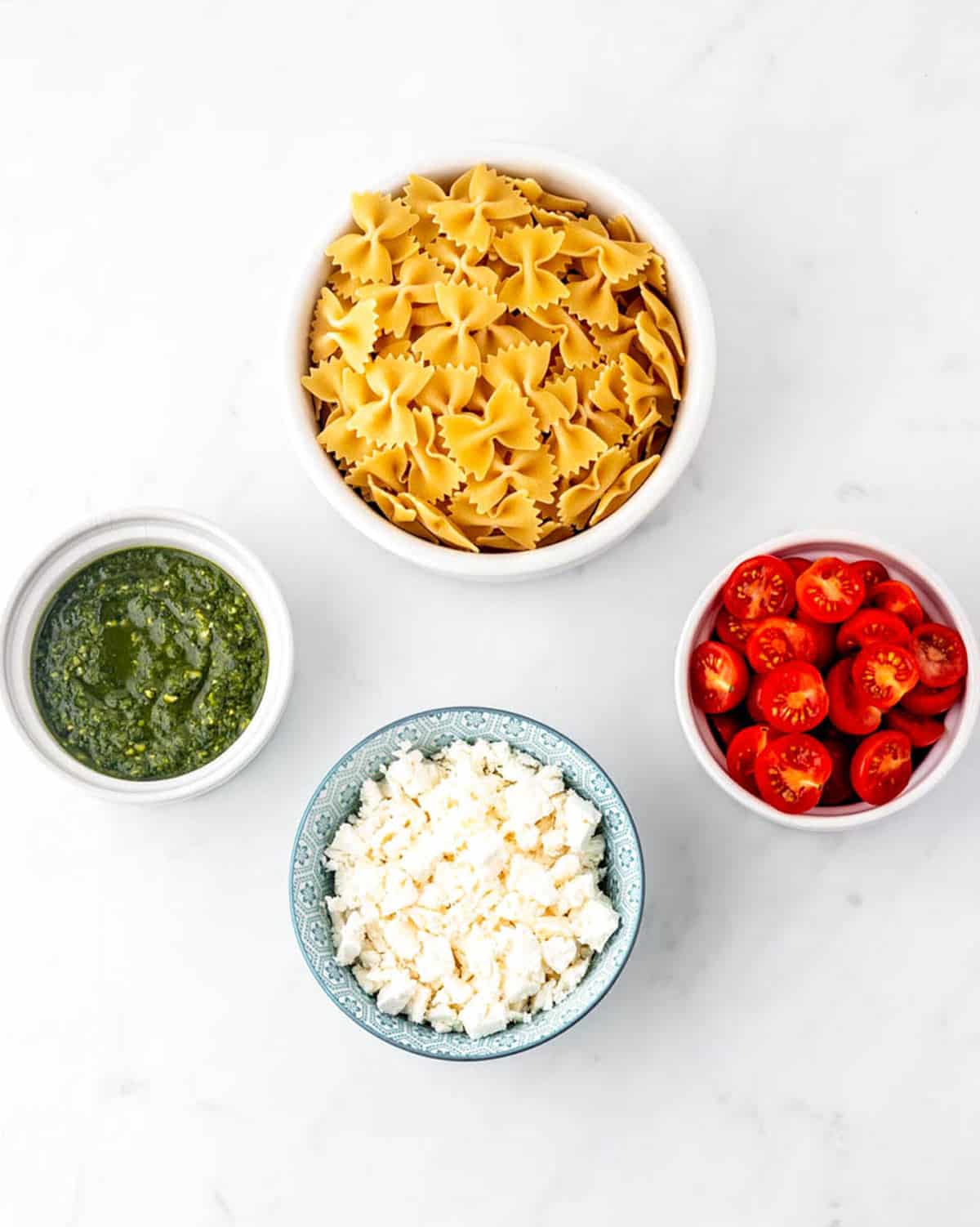 Bowtie pasta, tomatoes, feta cheese and pesto in bowls for the 4 ingredient pasta salad recipe.