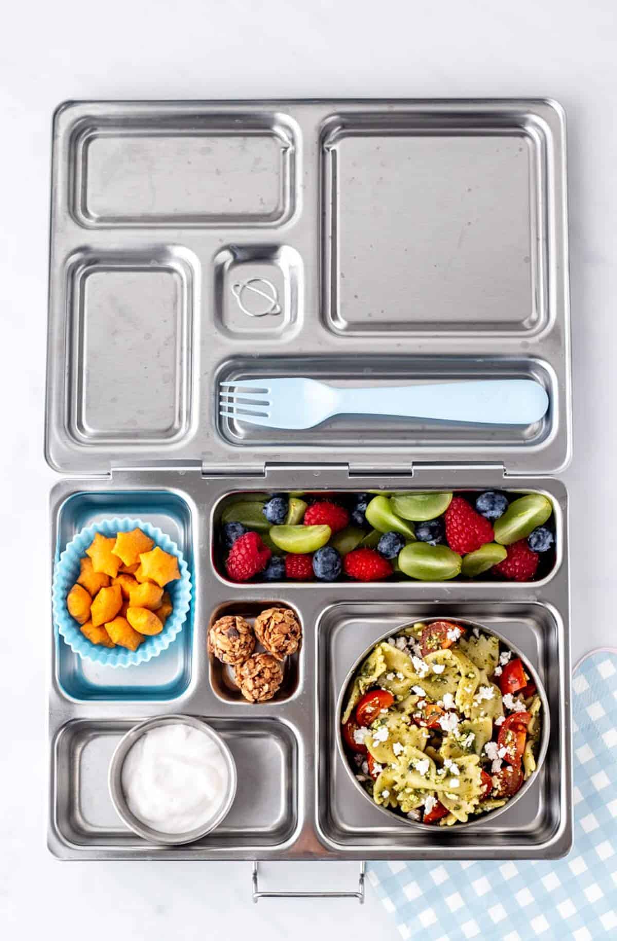 A stainless steel lunch box with 4 ingredient pasta salad, fruit, star crackers, yogurt and granola bites.