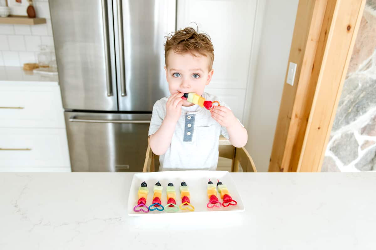 A toddler eating fruit off a skewer.