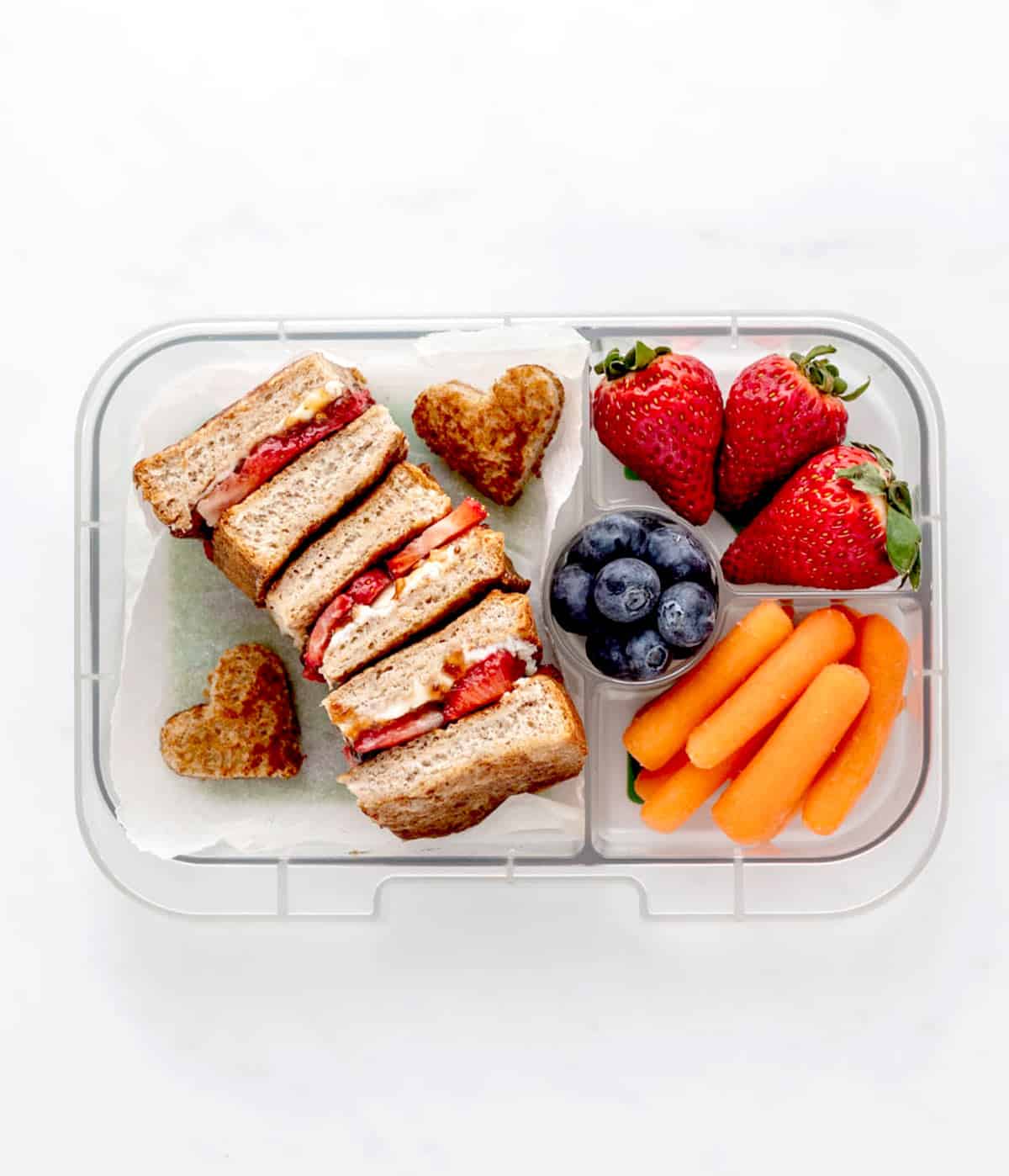 A strawberry cream cheese banana French toast sandwich in a lunch box with strawberries, blueberries and carrot sticks.