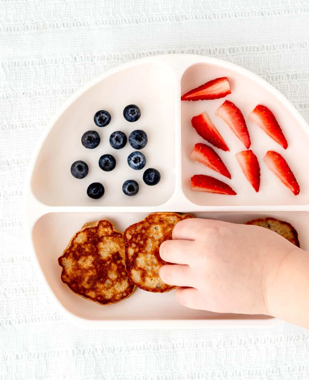 A toddler's hand picking up a banana oat pancake off of a white plate with blueberries and strawberries.