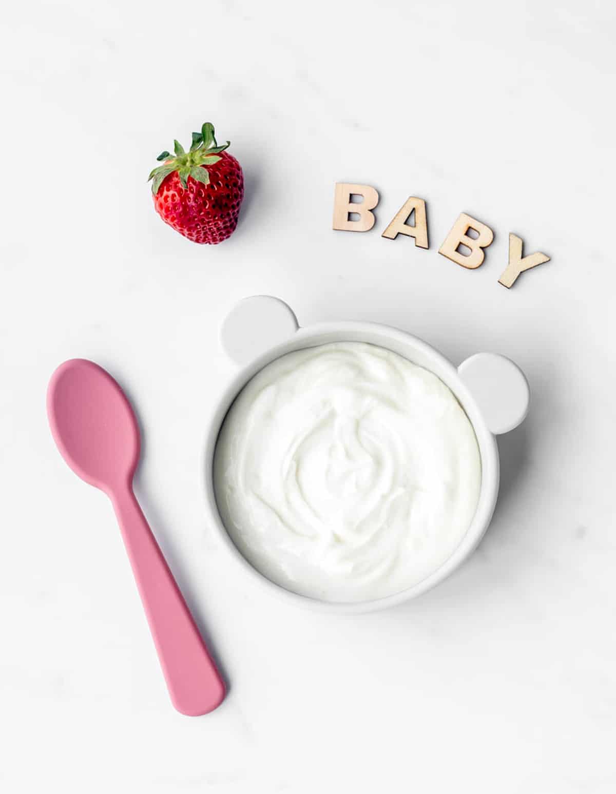 A bowl of yogurt next to a spoon, strawberry and word baby spelled out with wooden letters.