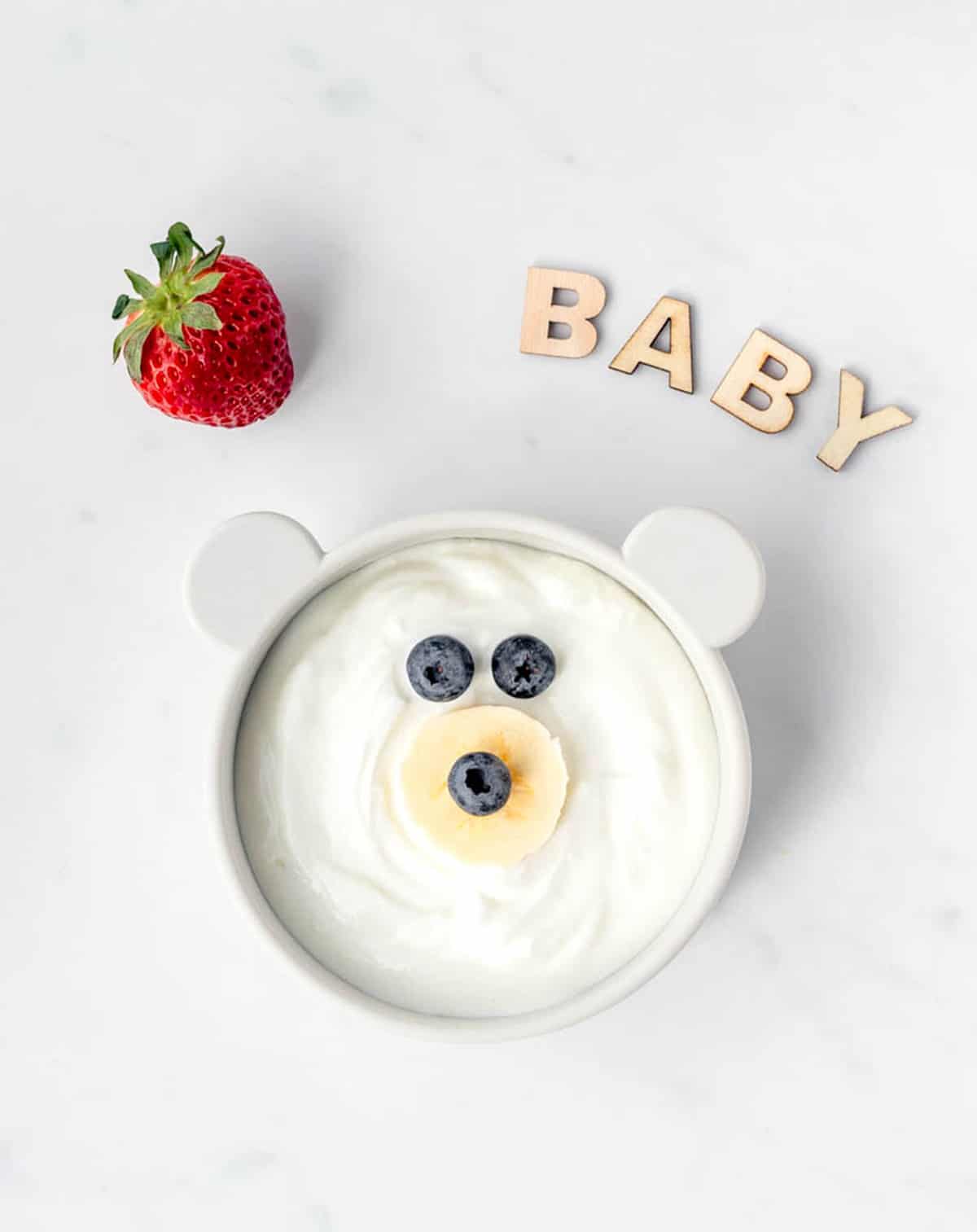 A teddy bear bowl filled with yogurt with a strawberry and word baby spelled out next to it.