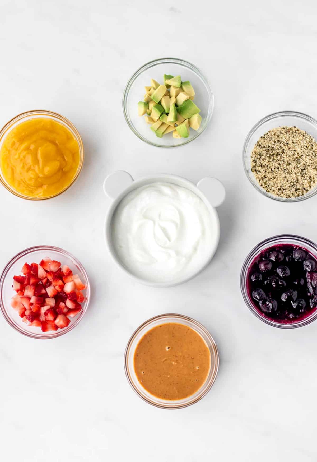 A bowl of yogurt surrounded by bowls of fruit, nut butter, avocado and hemp hearts.