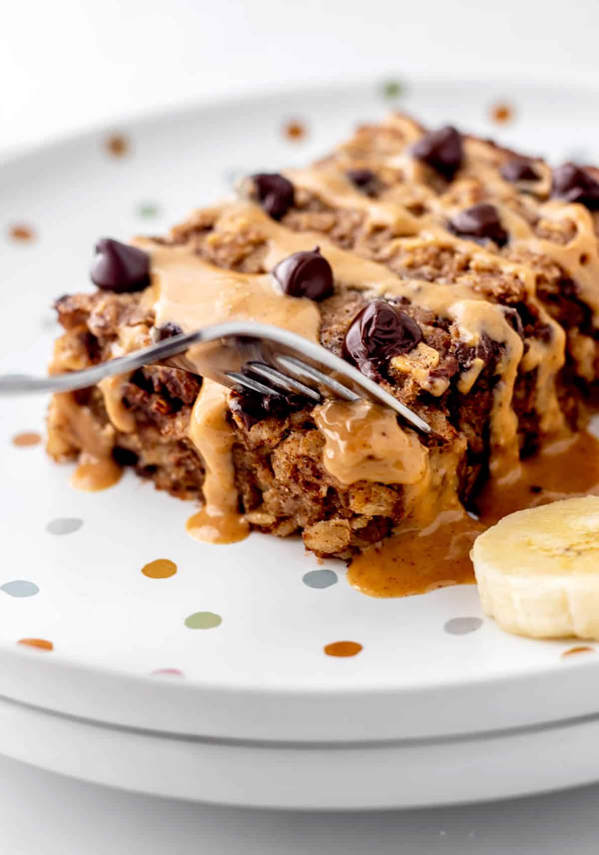A fork digging into a piece of baked oatmeal with chocolate chips, topped with a drizzle of peanut butter.