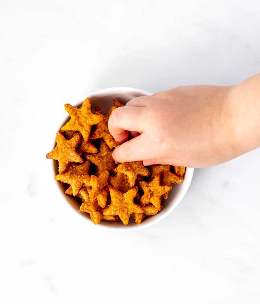 A child's hand picking up a sweet potato cracker out of a white bowl.