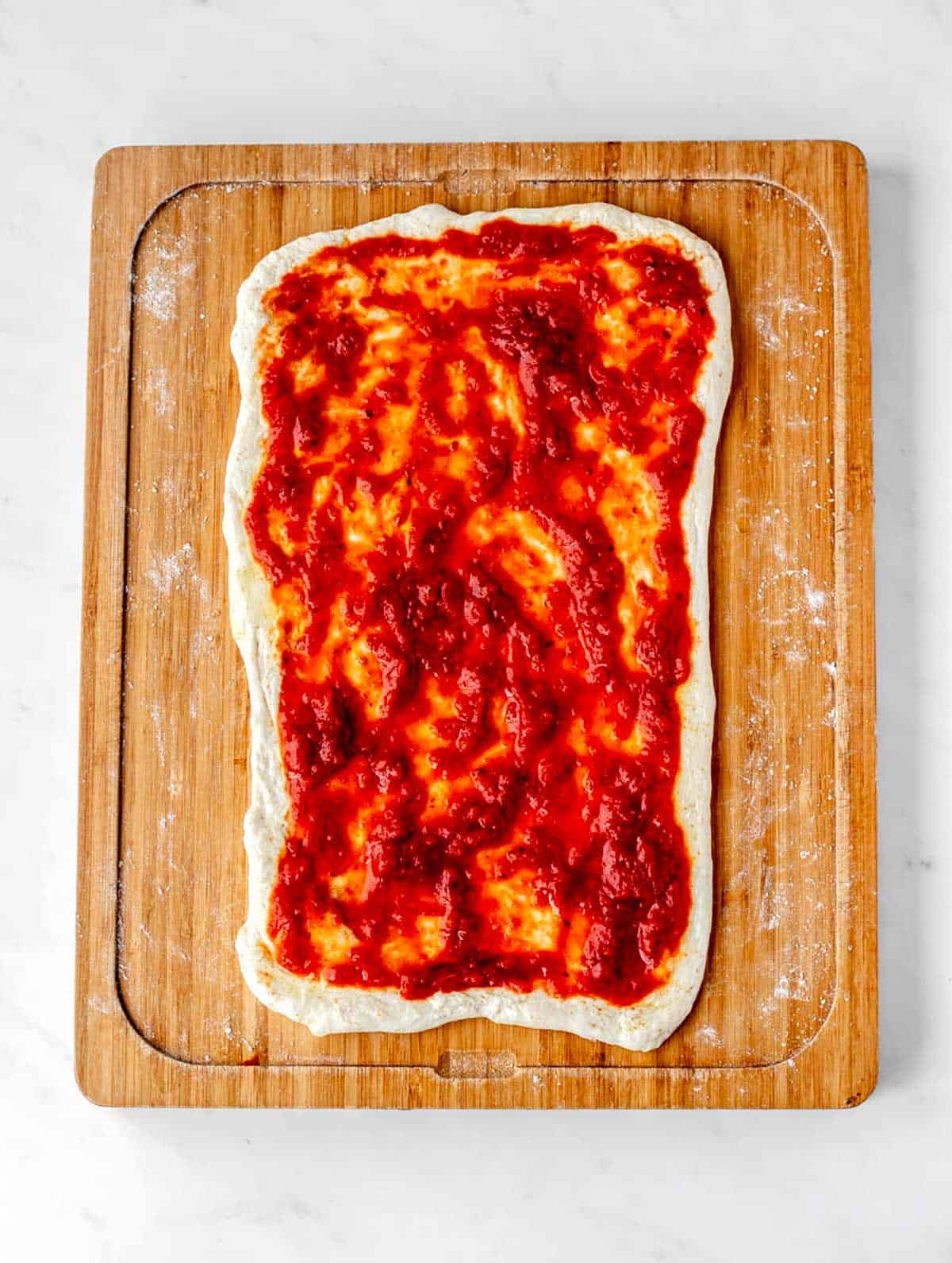 A layer of pizza sauce on top of the rolled out dough on a cutting board.