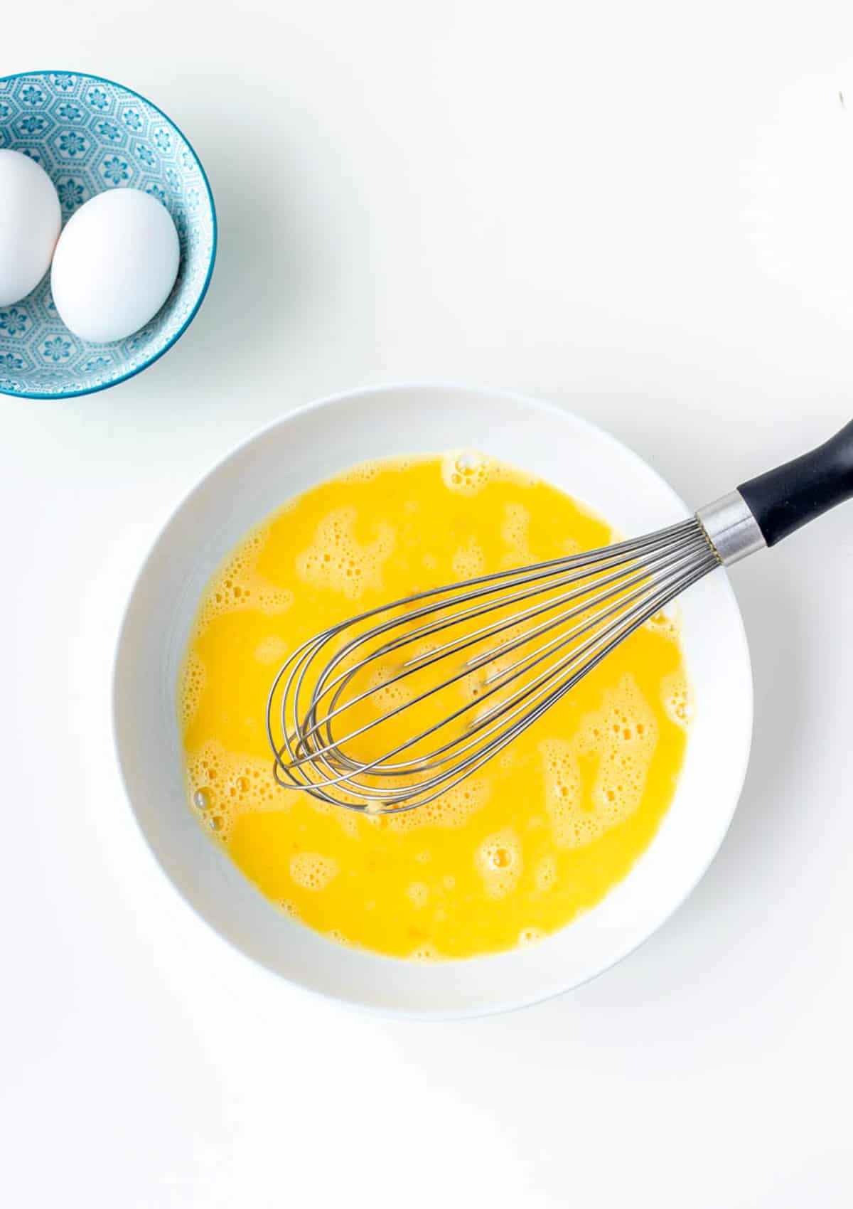 A whisk in a white bowl with egg mixture to make scrambled eggs.