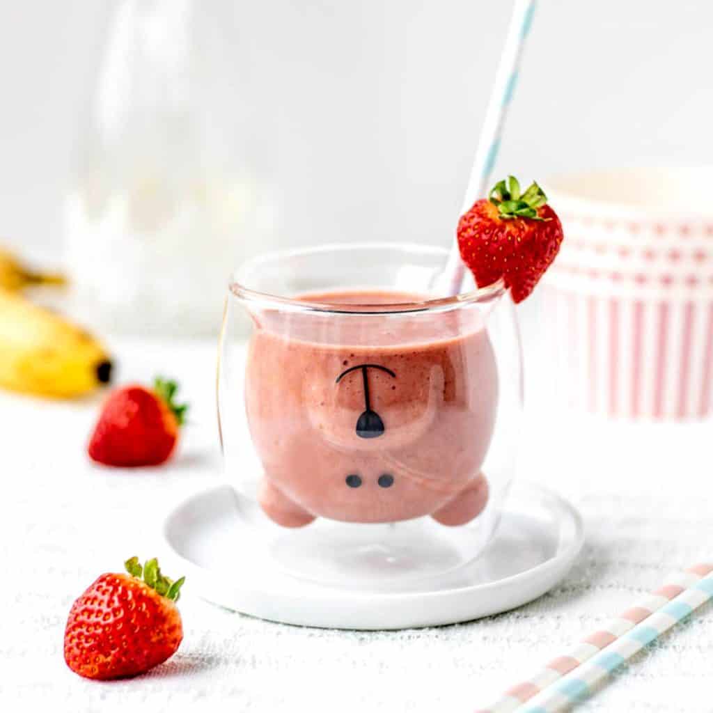 A teddy bear glass filled with strawberry banana smoothie with a fresh strawberries.