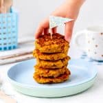 A sweet potato patty being picked up by a child's hand off of a stack of fritters.