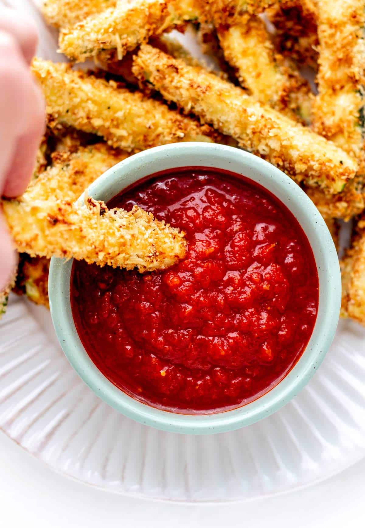 A hand dipping a zucchini fry into a bowl of marinara.