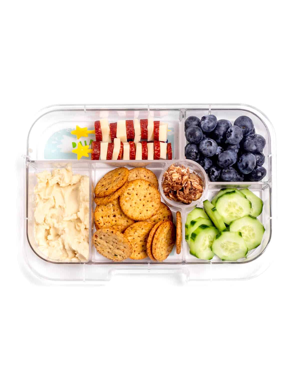 A lunch box with hummus, whole grain crackers, granola bites, cucumber slices, blueberries and mini turkey and pepperoni cheese skewers.