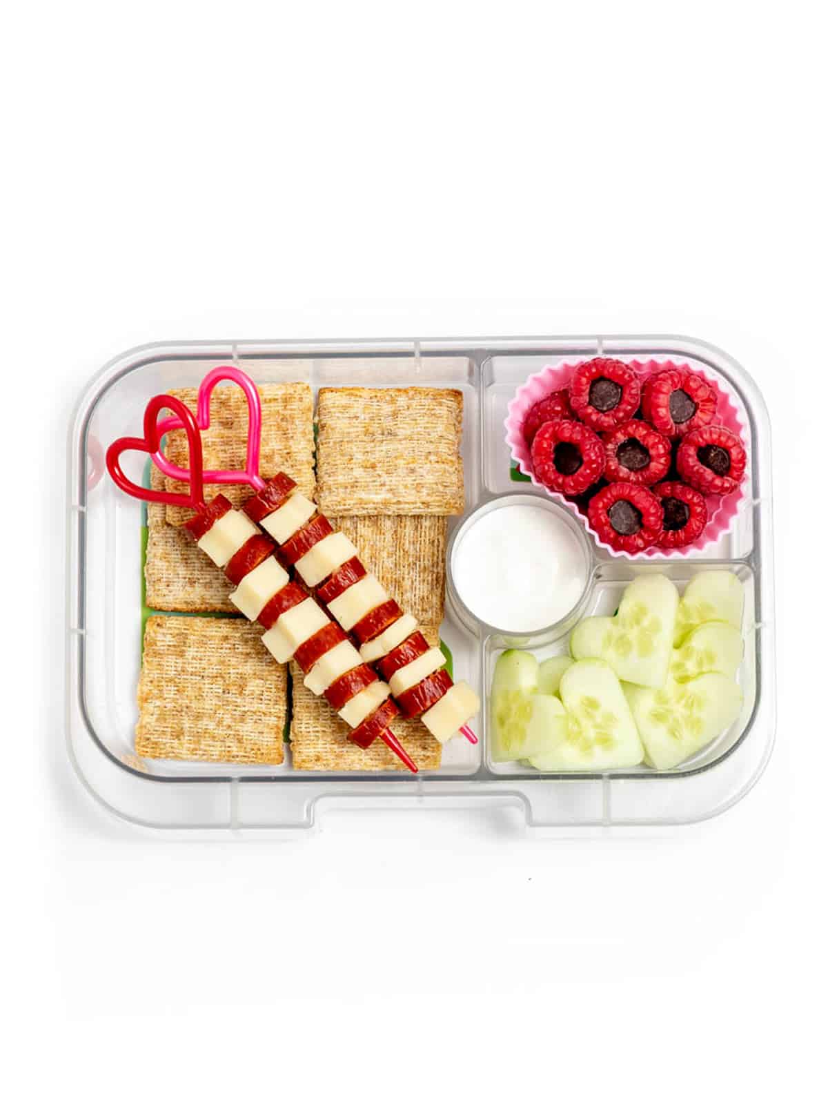A lunch box with two heart skewers with cheese cubes and mini pepperoni, triscuits, cucumber hearts, and dark chocolate chip stuffed raspberries.
