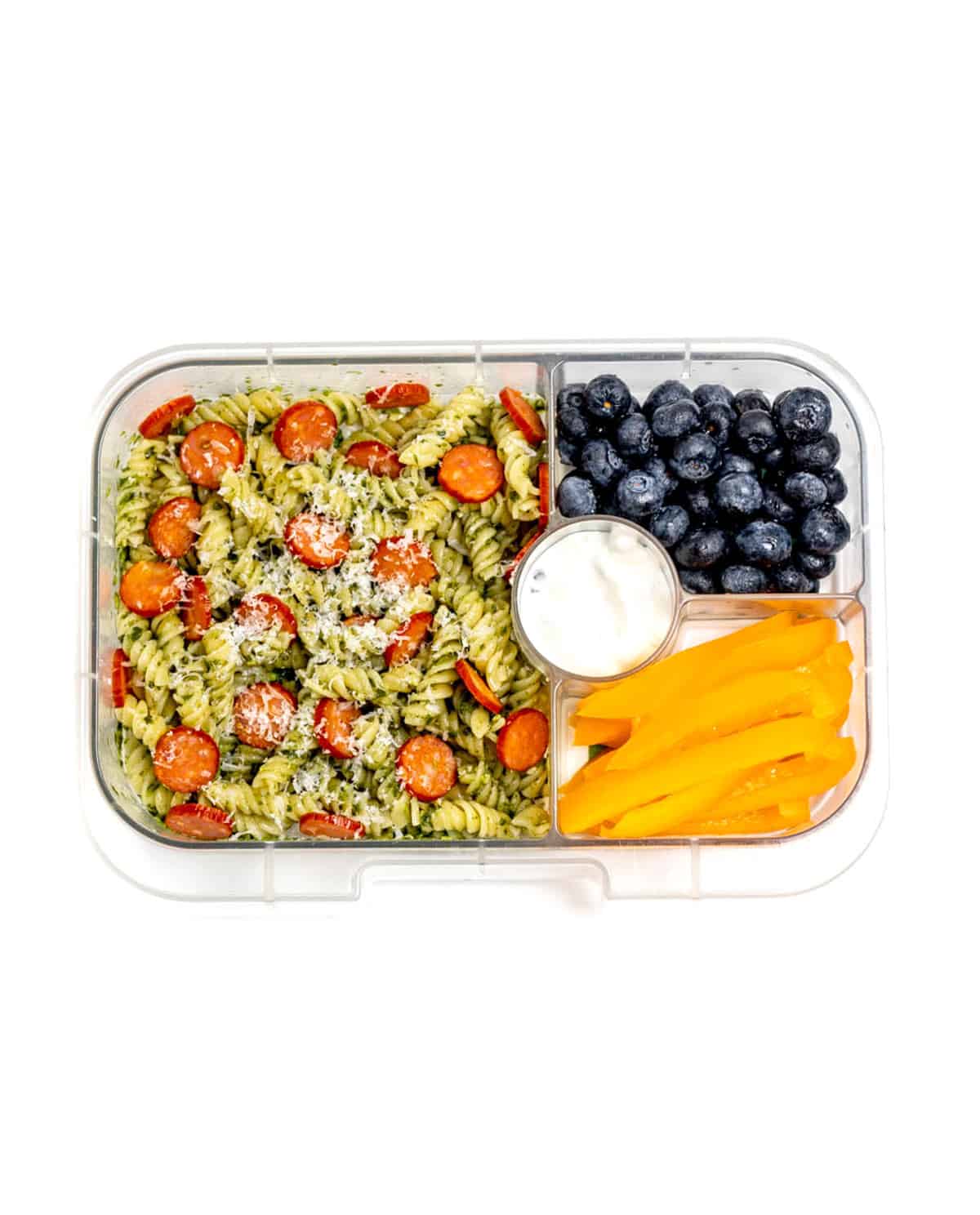 A lunch box with pesto pasta salad with mini pepperoni slices, parmesan cheese, served with yellow pepper slices, dip and blueberries.