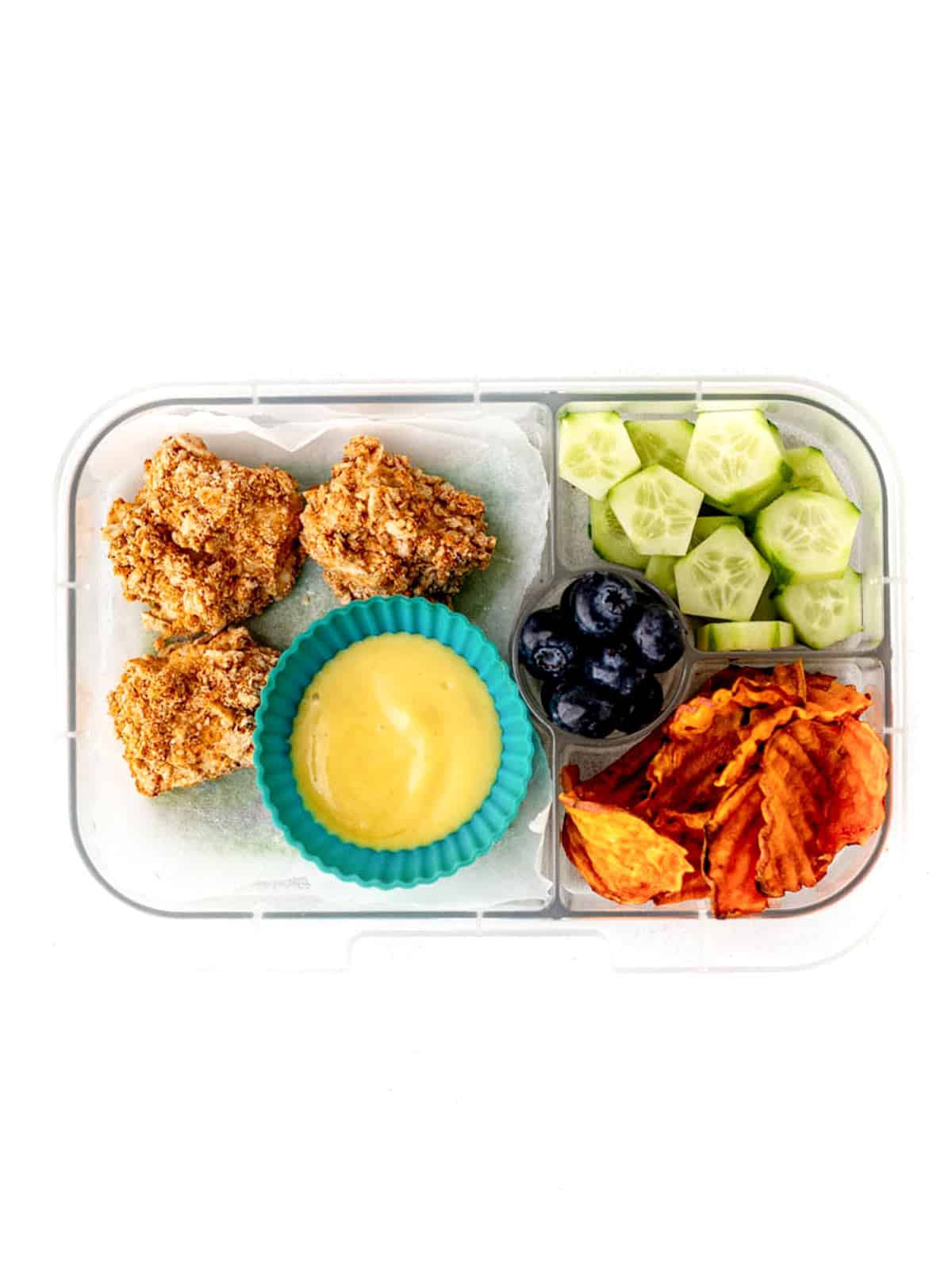 A lunch box with leftover air fryer chicken bites, honey mustard dip, baked sweet potato chips, cucumber slices and blueberries.