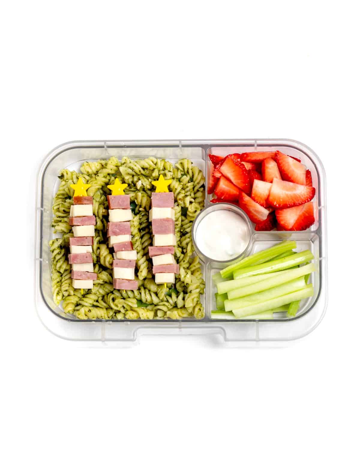 A lunch box with pesto pasta salad, topped with star ham and cheese skewers, strawberries, celery sticks and dip.