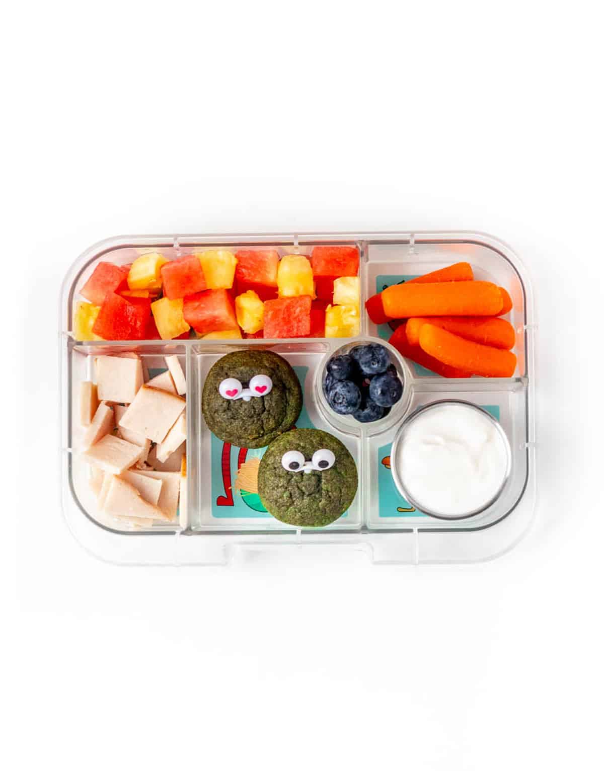 A lunch box with deli turkey squares, green monster muffins, watermelon, pineapple, blueberries, carrot sticks and Greek yogurt.