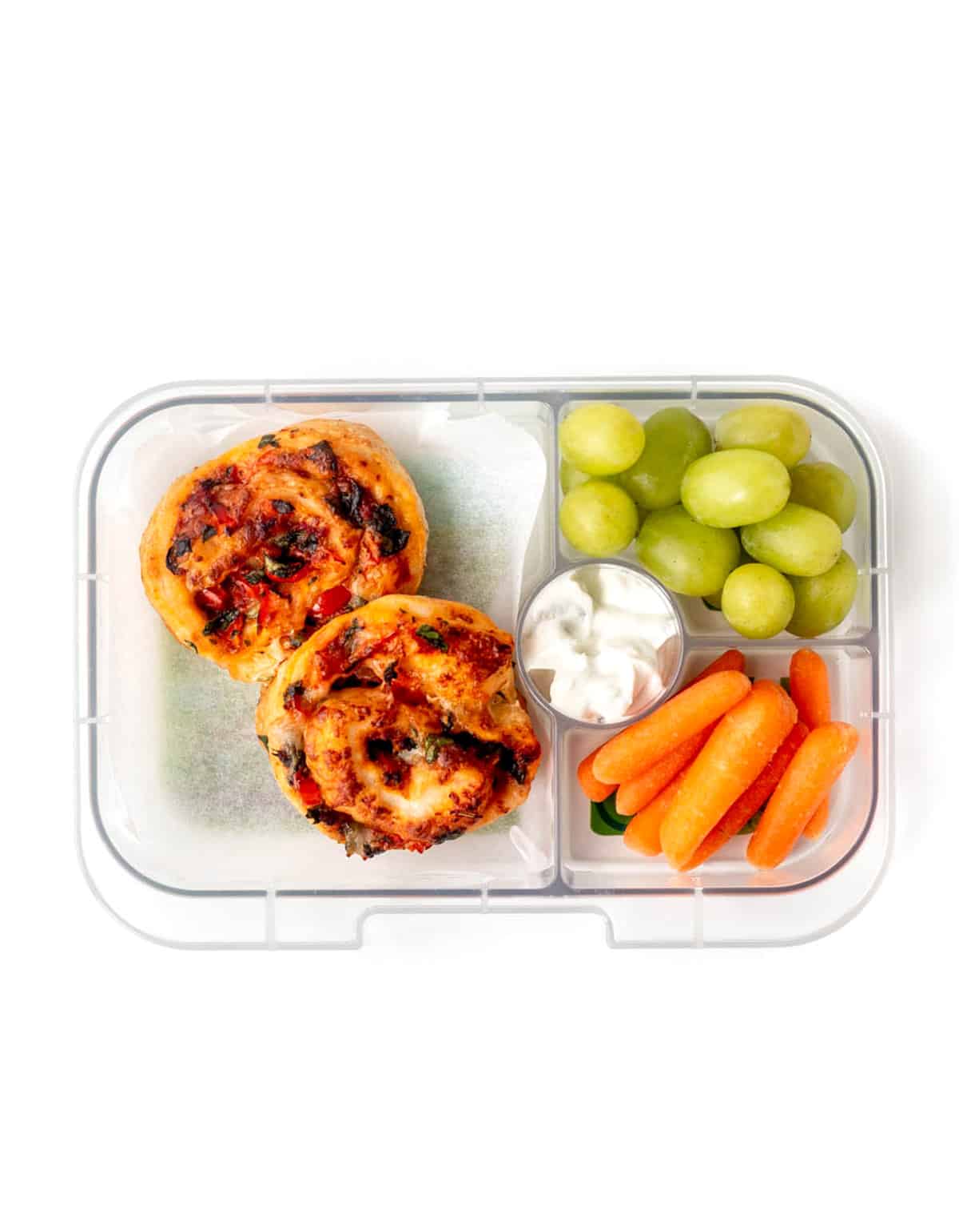 A lunch box with two veggie pizza rolls, grapes, carrots and tzatziki.