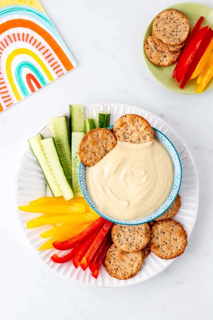 Two pita crackers dipped in a bowl of high protein hummus on a plate of veggies and crackers.