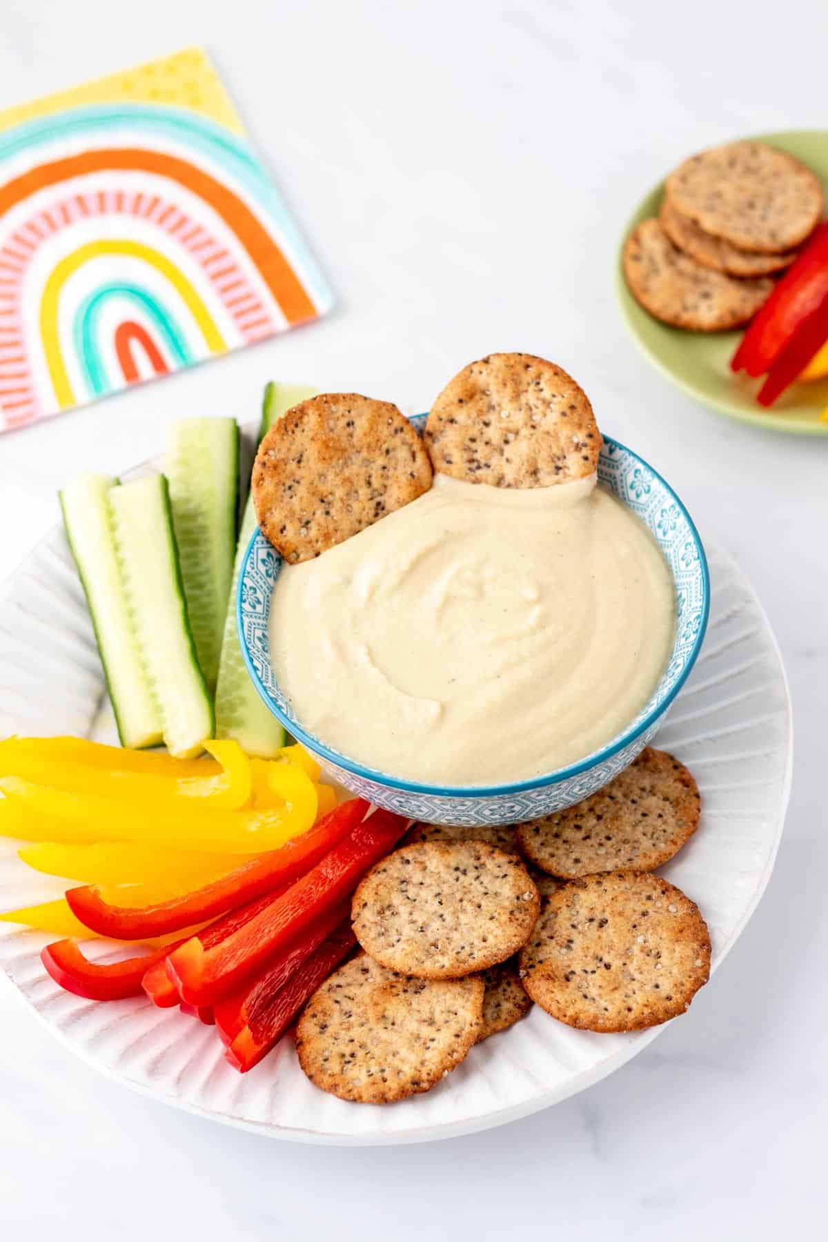 A white plate with a bowl of high protein hummus with two crackers dipped in it.