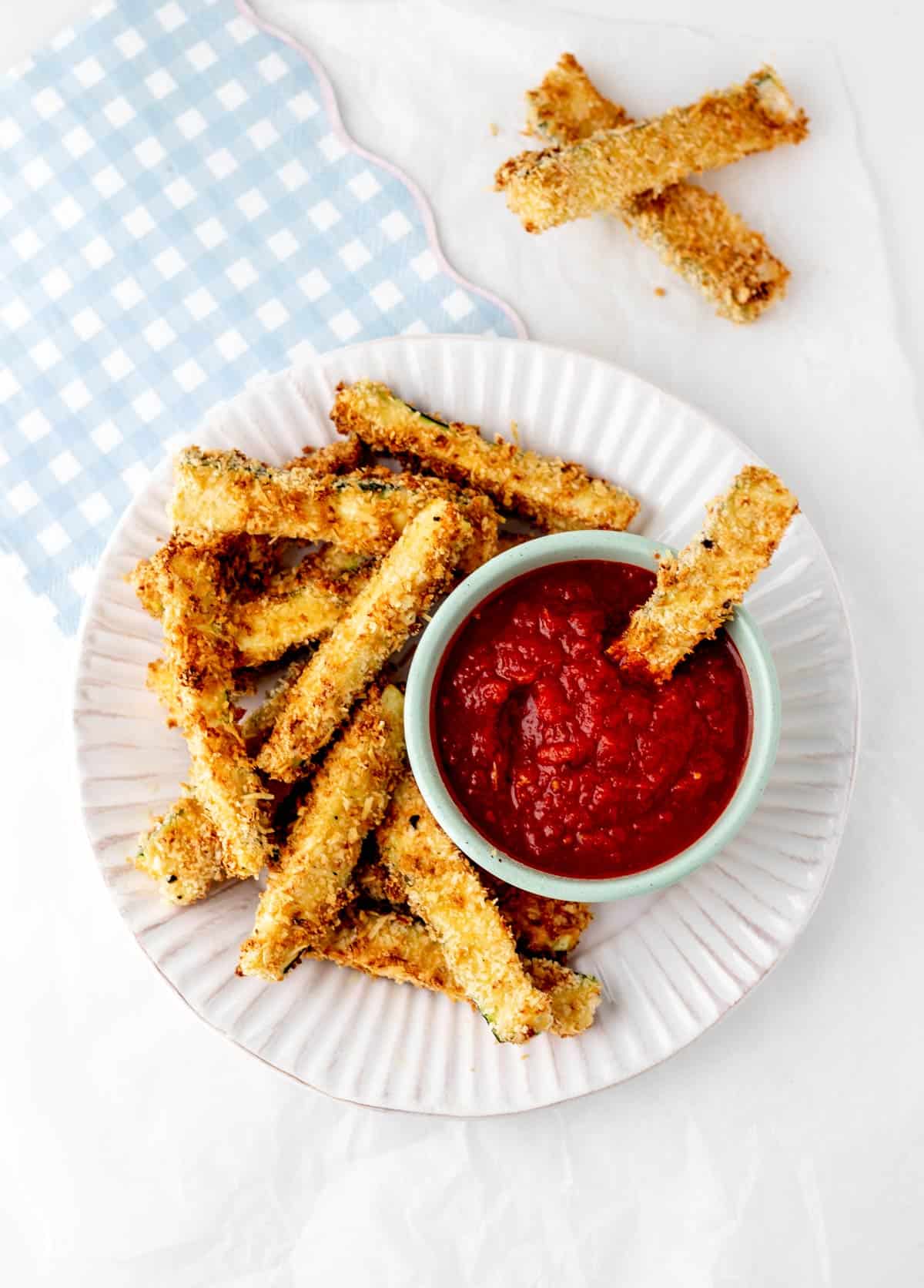An air fryer zucchini fry dipped in a bowl of marinara on a plate of fries.