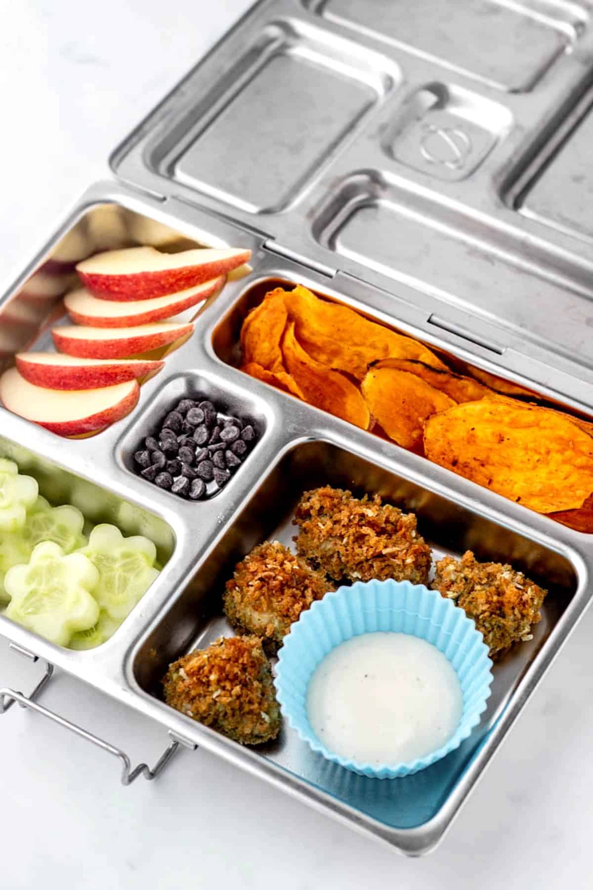 Pesto chicken bites in a lunch box with apples, cucumbers, sweet potato chips and chocolate chips.