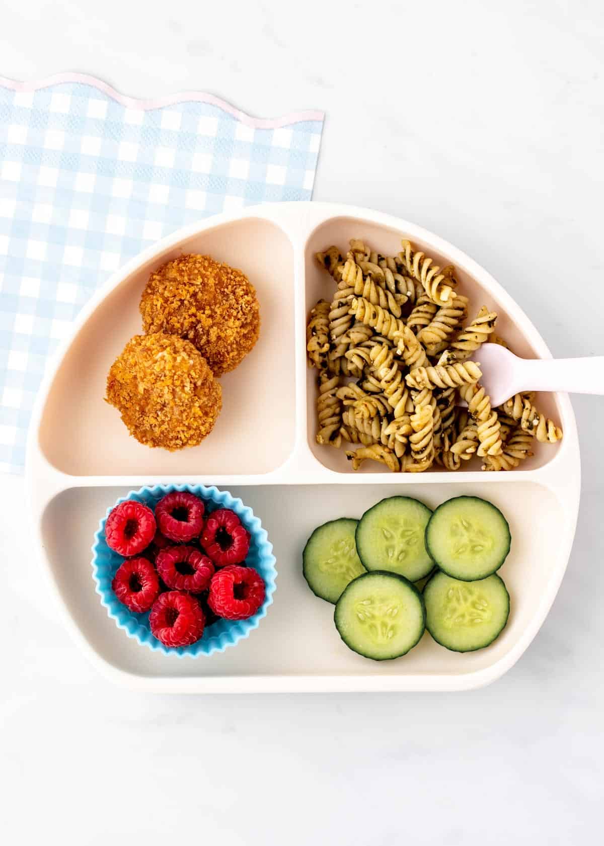A divided plate with chicken nuggets, noodles, raspberries and cucumbers.