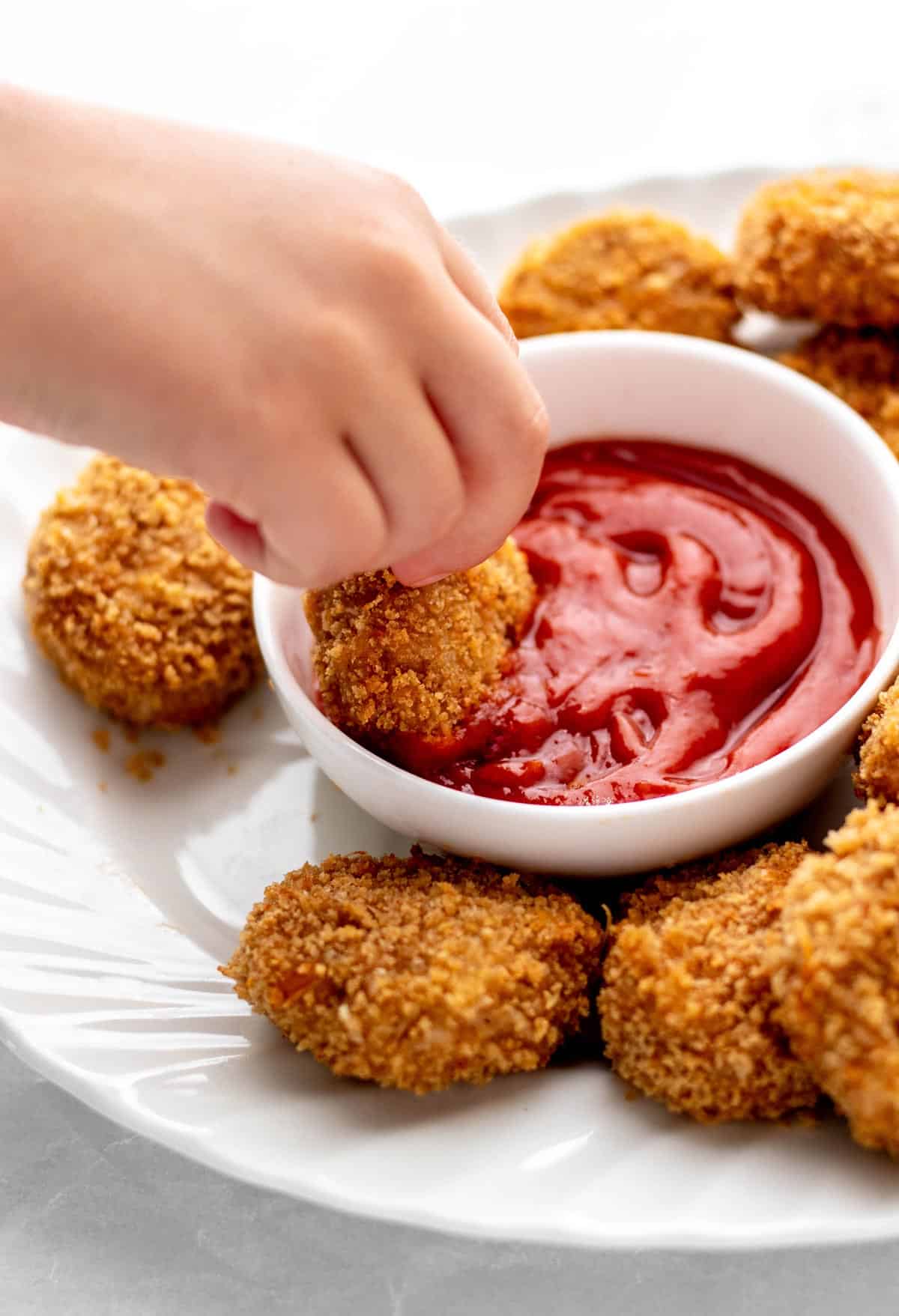 A child's hand dipping a chicken nugget into a bowl of ketchup.