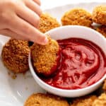 A child's hand dipping a crispy ground chicken nugget into a bowl of ketchup.