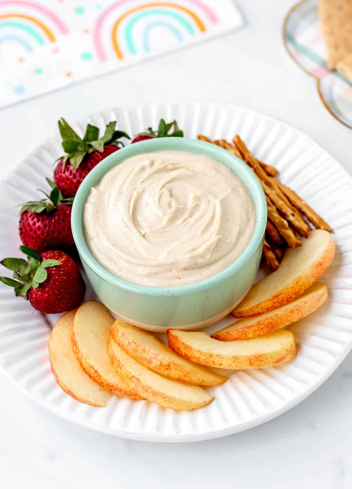 A small bowl of peanut butter fruit dip on a plate with fruit and pretzels.