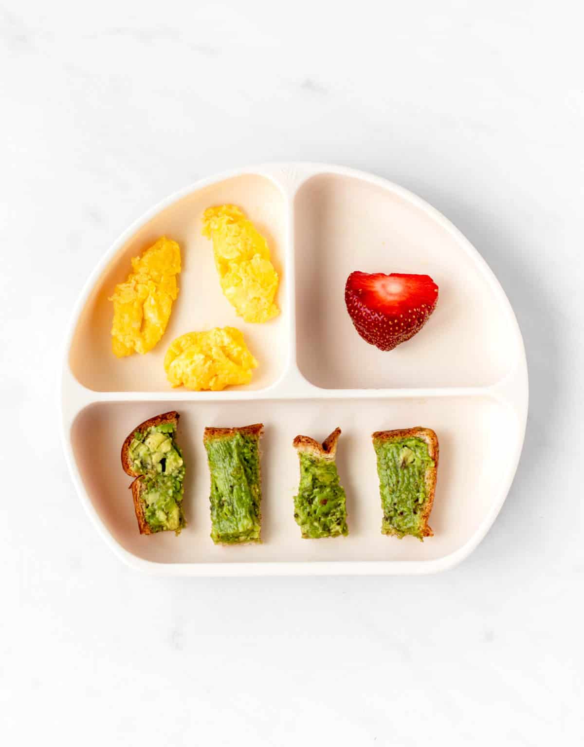 Scrambled eggs, avocado toast strips and a strawberry on a divided plate.