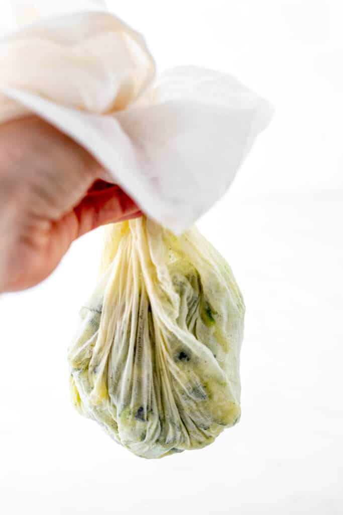 A hand holding a cheesecloth with ringed out zucchini.