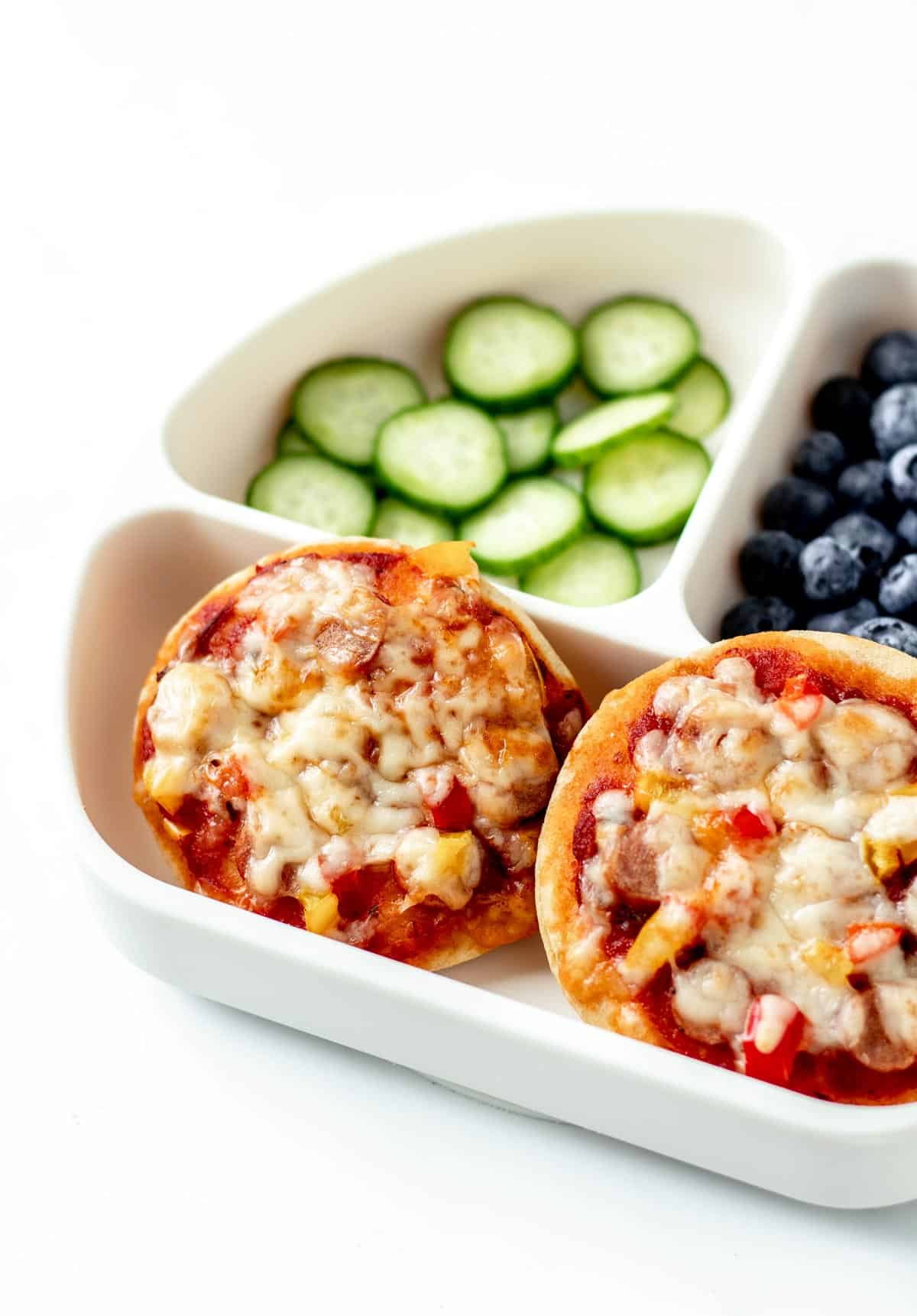 Two homemade mini pizzas on a plate with cucumbers and blueberries.