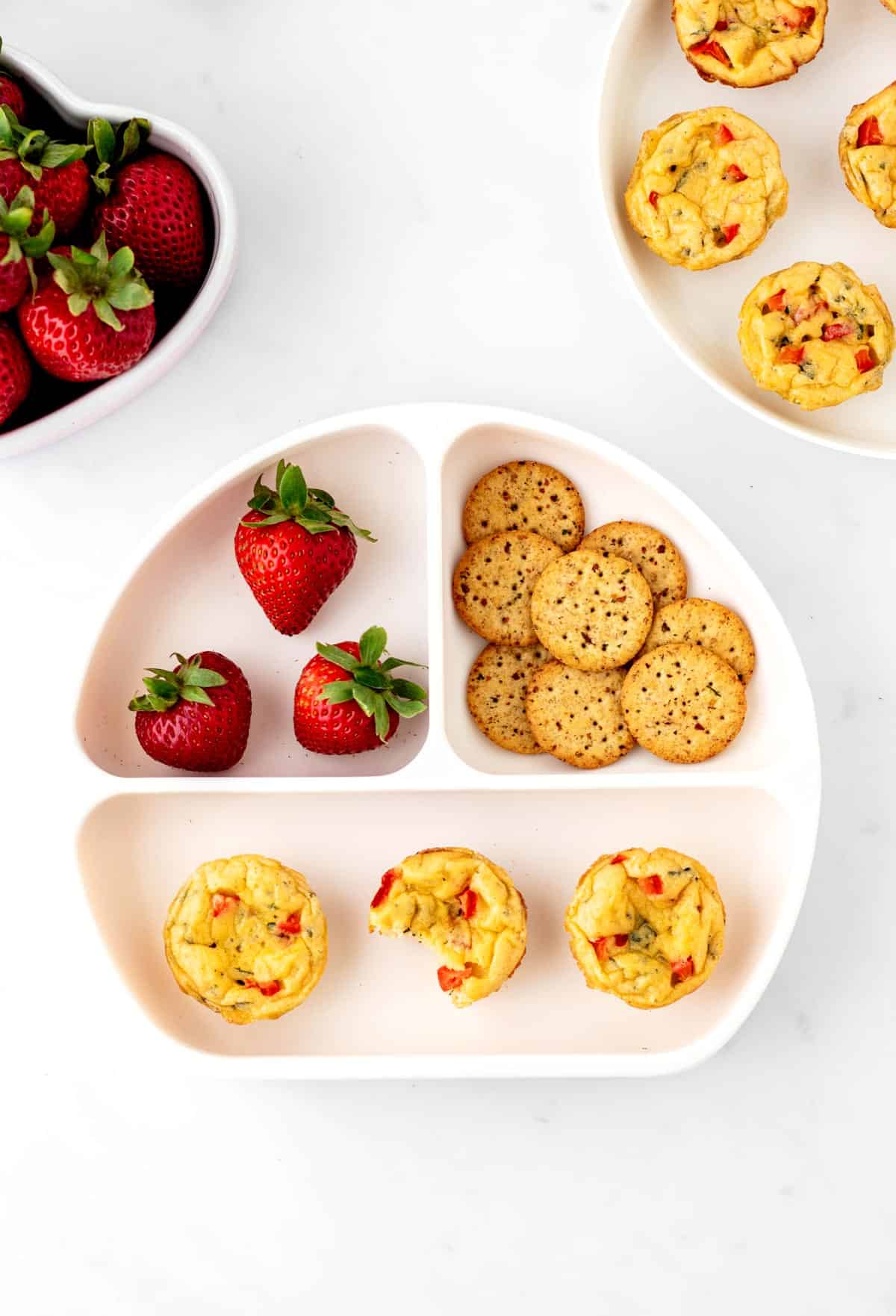 Three egg bites on a divided plate with crackers and strawberries.
