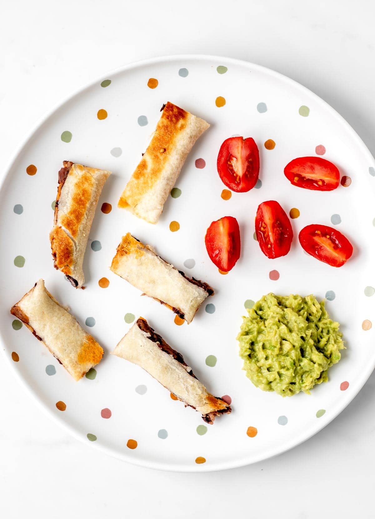 Pieces of a baby led weaning quesadilla, guacamole and tomatoes on a polka dot plate.