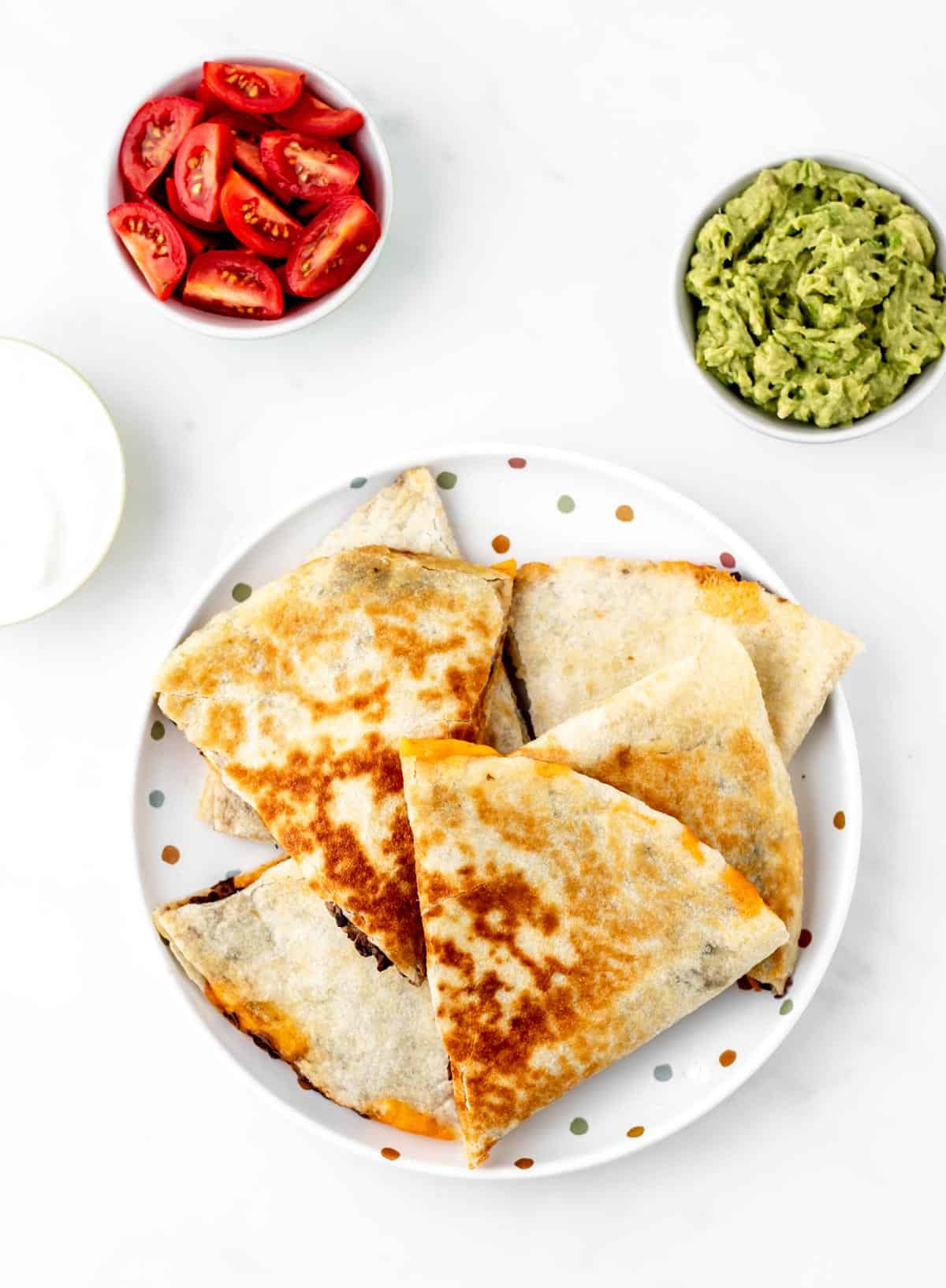 A pile of baby lead weaning quesadillas on a polka dot plate with guacamole and tomatoes.