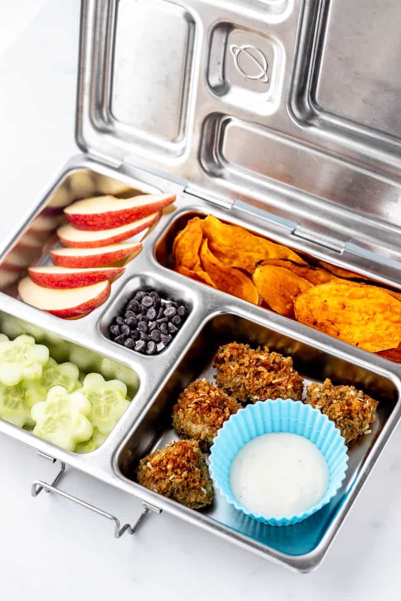 A stainless steel lunch box with chicken nuggets, cucumber flowers, sweet potato chips, apple slices and chocolate chips.