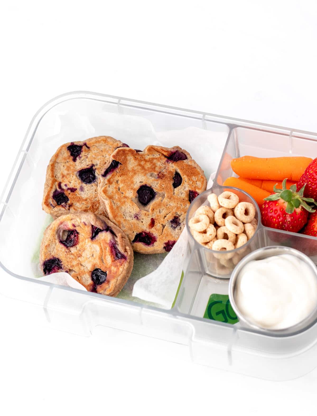 Mini blueberry pancakes in a lunch box with cheerios, yogurt, carrots and strawberries.
