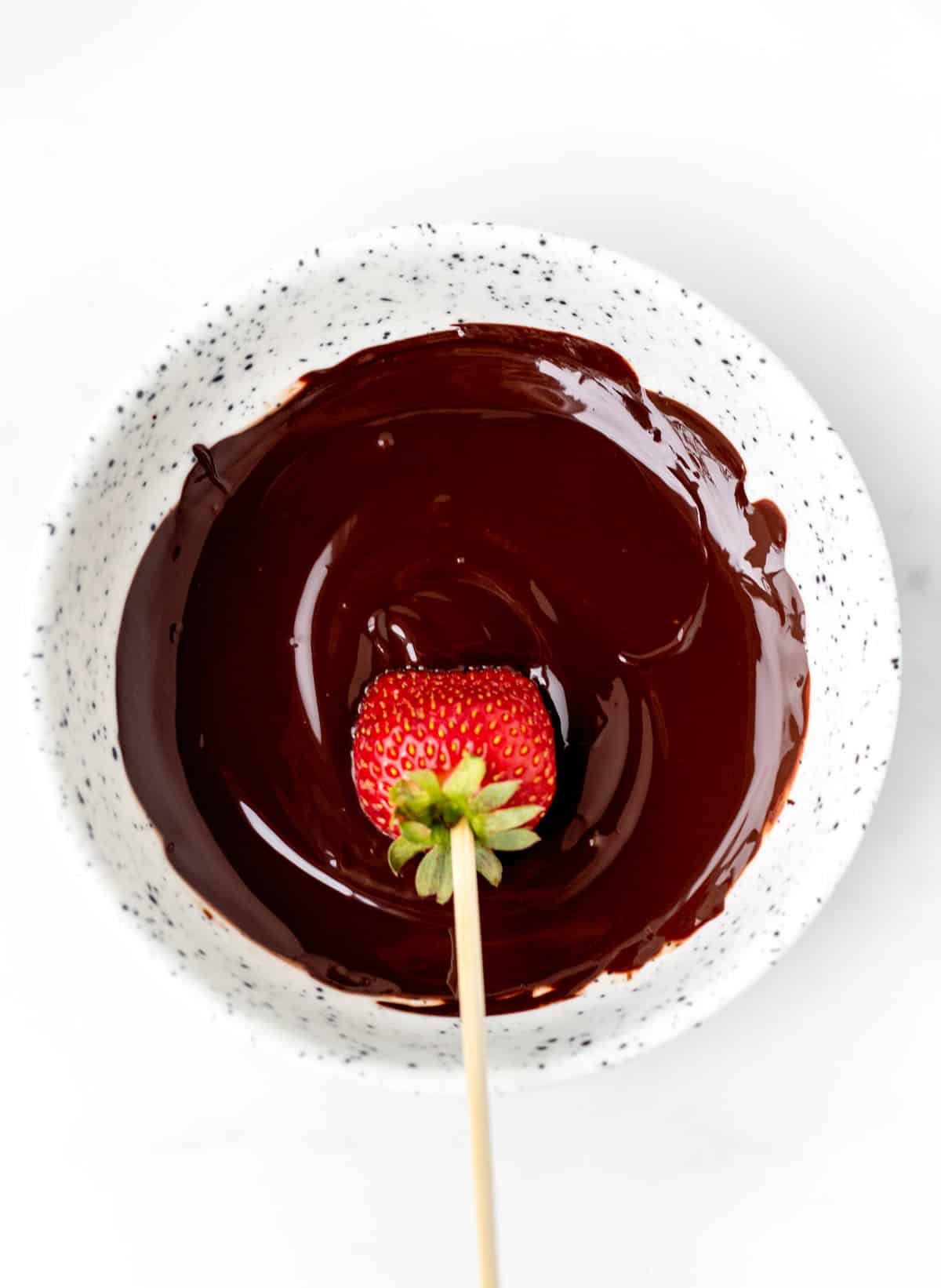A strawberry on a wooden skewer being dipped into a bowl of melted chocolate.