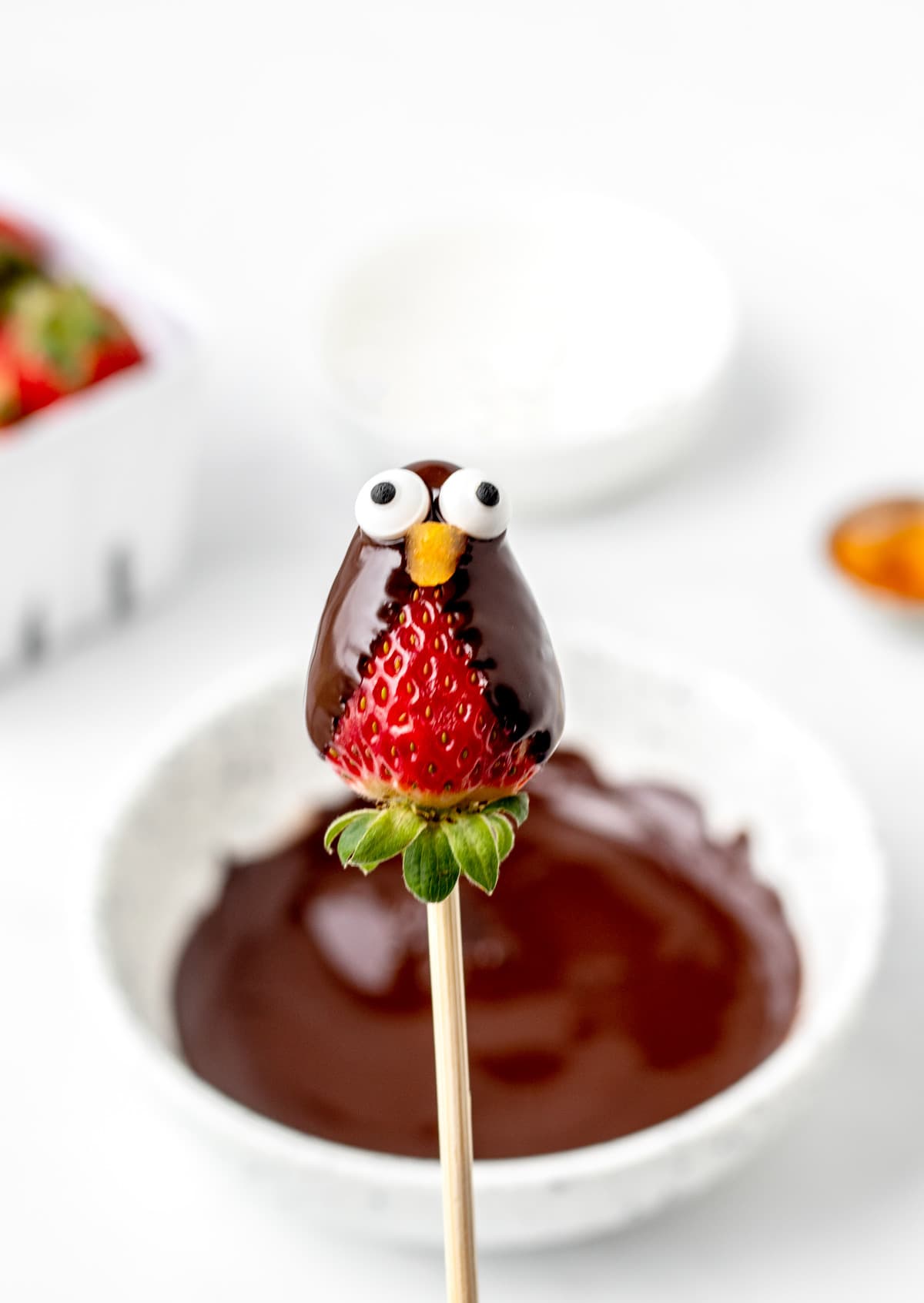 A chocolate covered strawberry on a wooden skewer with the candy eyes and dried apricot beak.