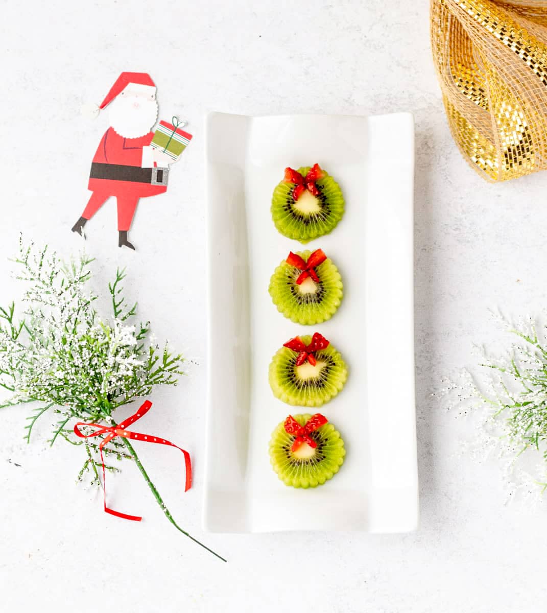 Four kiwi wreaths on a decorative white platter, surrounded by festive holiday decorations.
