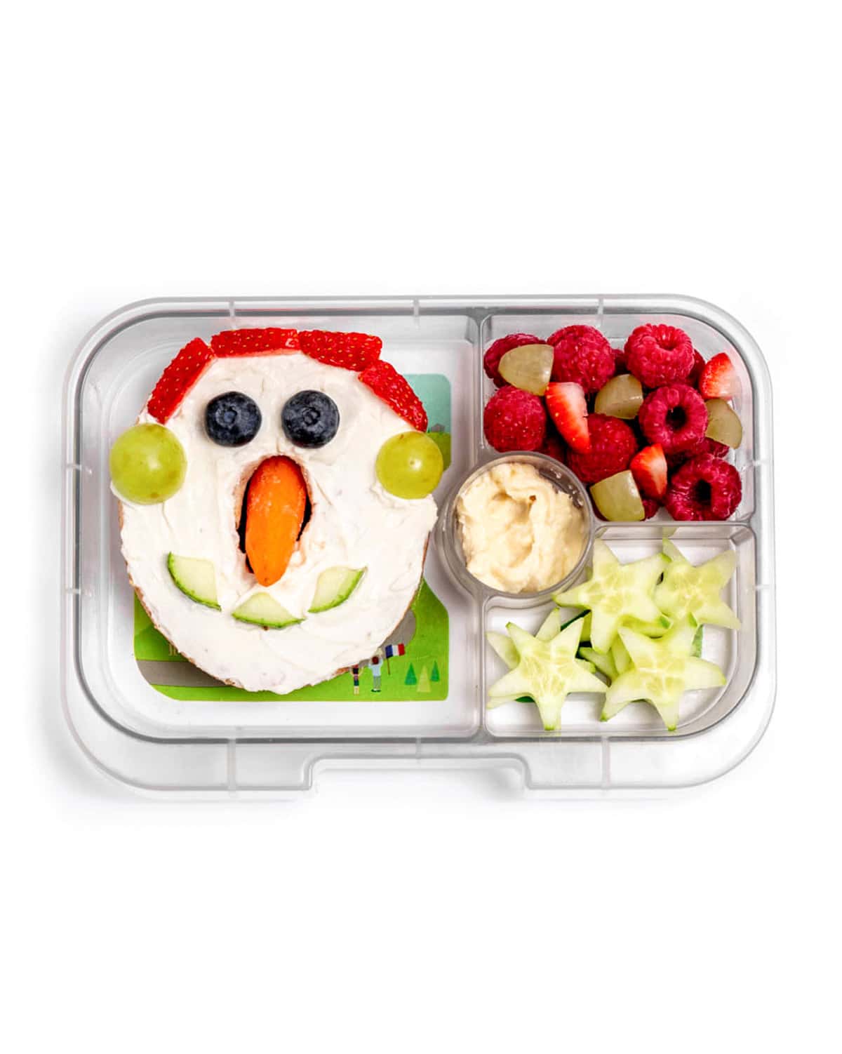 A lunchbox containing one snowman bagel, cucumber stars with a side of hummus, and a fruit mixtures of raspberries, strawberries and green grapes.