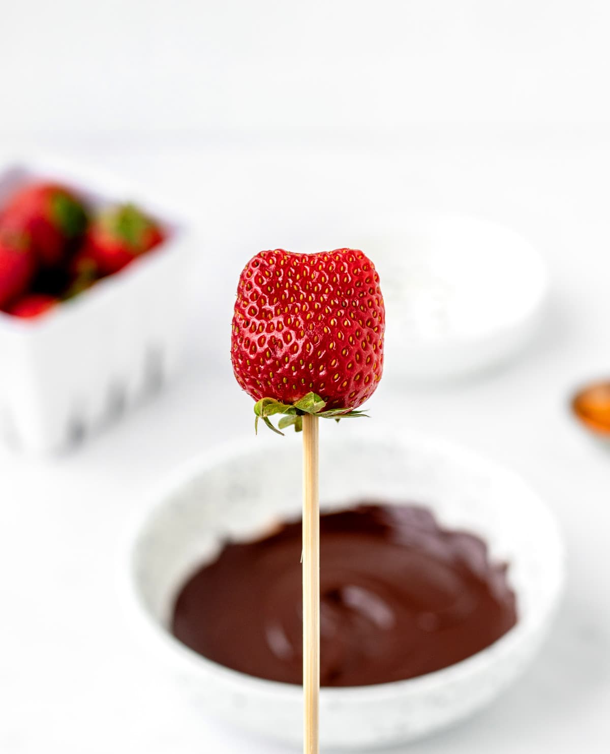 A strawberry on a wooden skewer with melted chocolate in white bowl in the background.