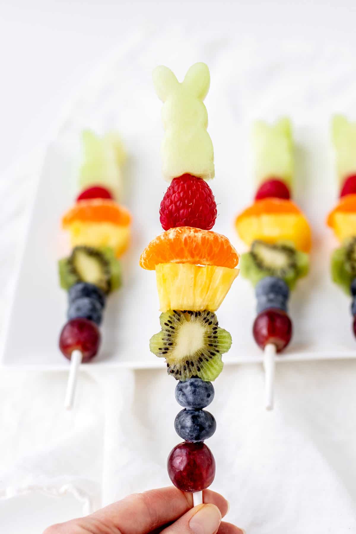 A close-up of someone holding an Easter fruit kabob with more fruit kabobs in the background on a white cutting board.