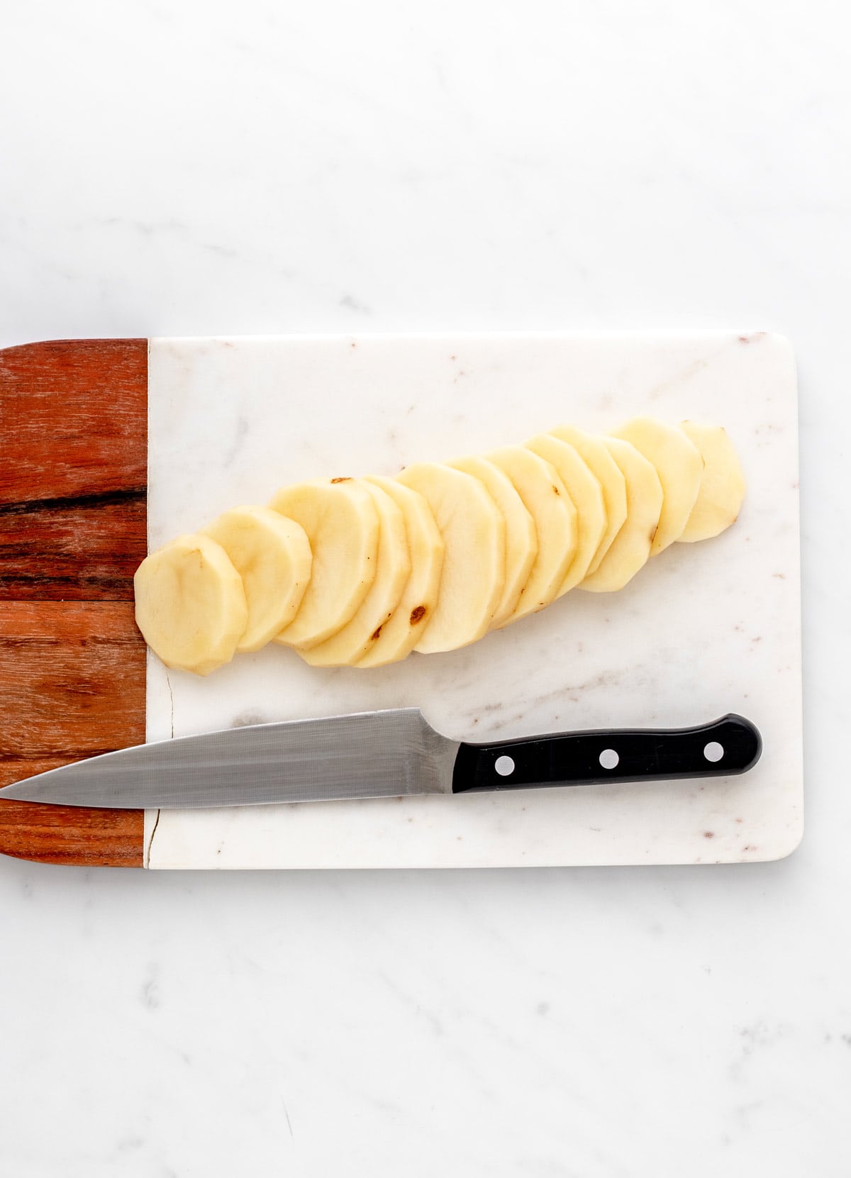 Russet potatoes cut into 1/4 inch slices on a wooden cutting board next to a knife.