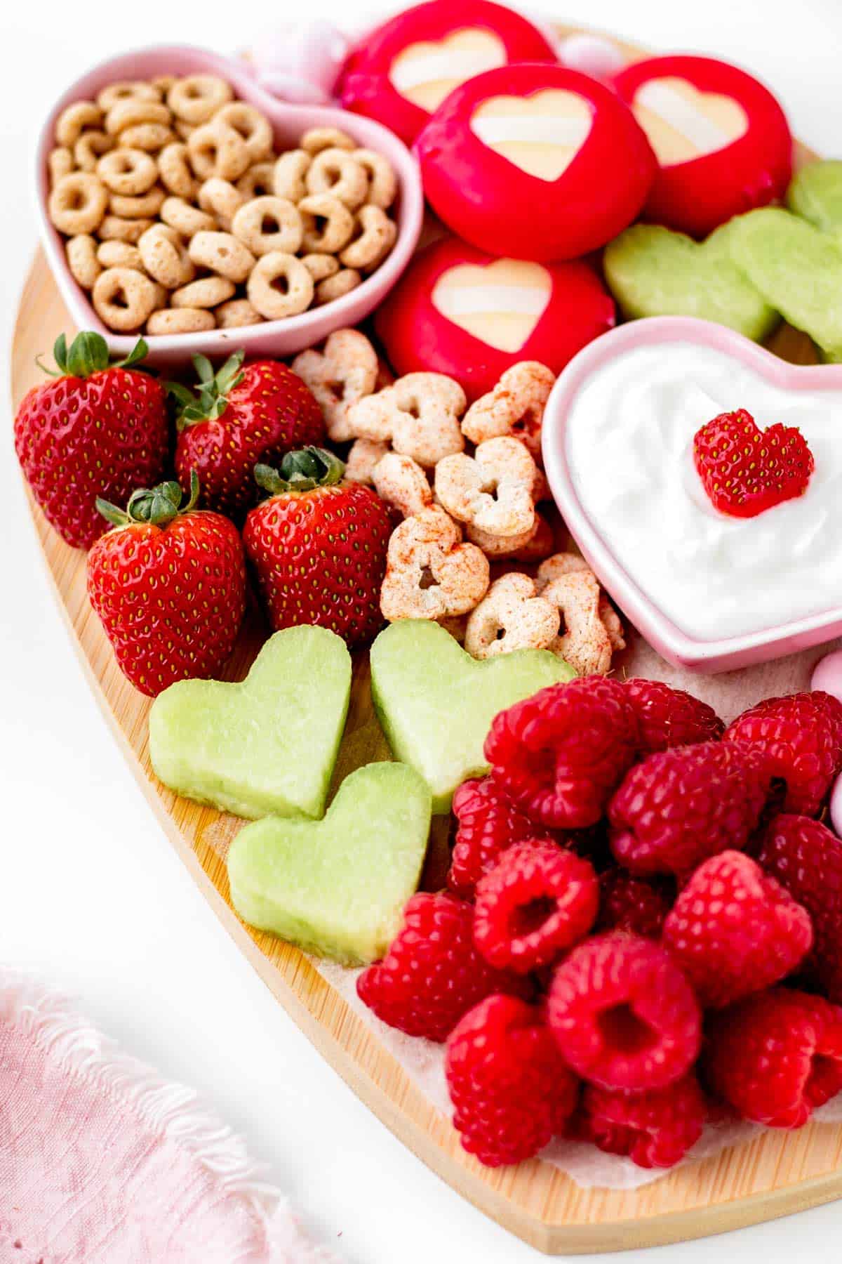 A close-up of the heart shaped charcuterie board showing raspberries, heart sandwiches, strawberries, yogurt with heart on top and cereal.