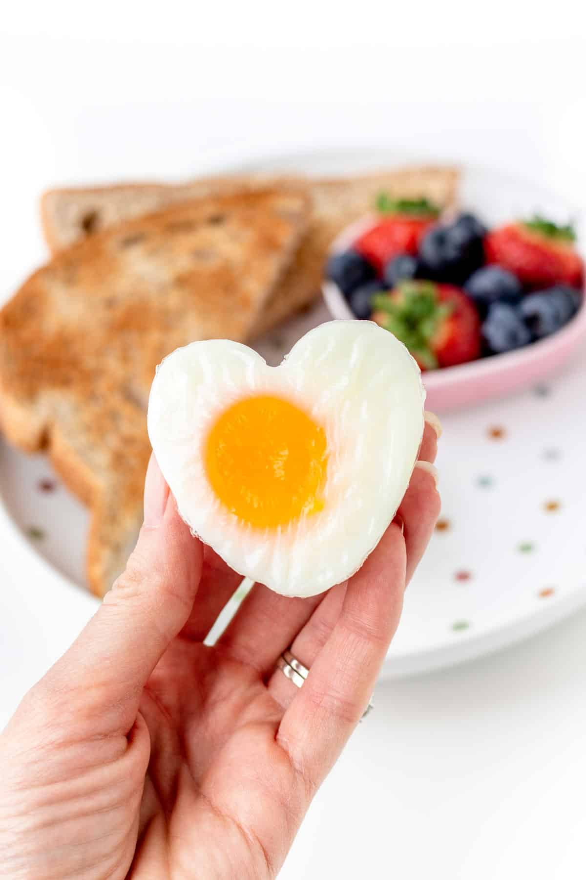 A close-up of a heart shaped egg in someone's hand with a plate of toast and berries in the background.