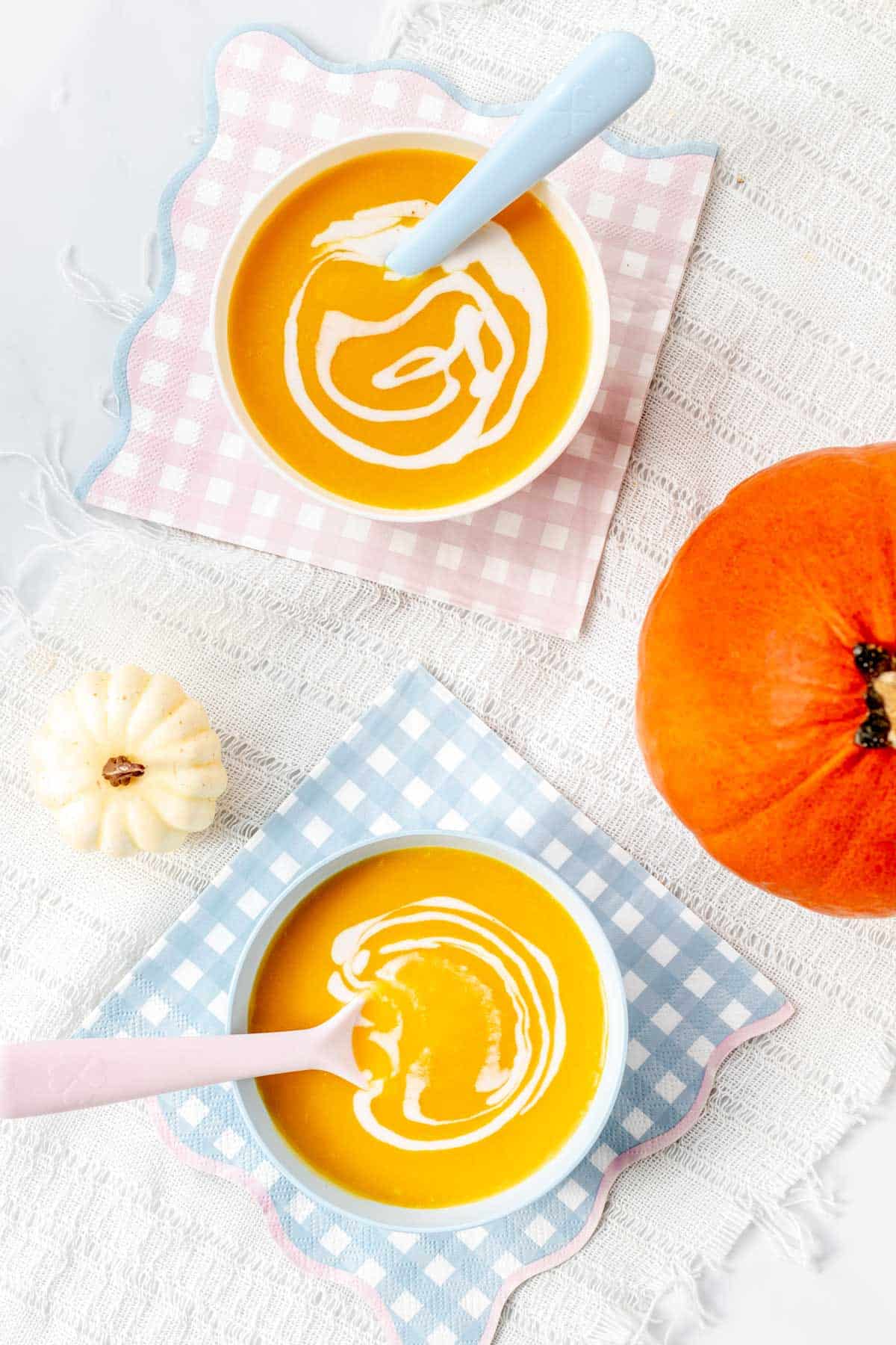 Two bowls of pumpkin soup with spoons in them on colorful napkins on a countertop, next to a pumpkin.