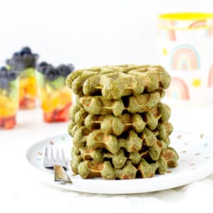A stack of spinach banana waffles on a plate next to rainbow fruit cups.